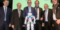 Sechs Männer in Anzügen, einer von ihnen hält einen kleinen weißen Roboter in Händen.