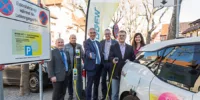 Eine Gruppe Menschen steht um eine Ladesäule und ein Elektroauto herum. Einige haben Ladekabel in der Hand.