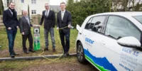 Vier Männer in Anzügen stehen naben einer Ladesäule und einem Elektroauto. Ein Mann hält ein Ladekabel in Händen.