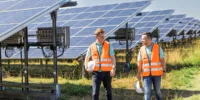 Zwei Männer mit Warnwesten spazieren an einem sonnigen Tag direkt neben einem Solarpark entlang