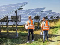 Zwei Männer mit Warnwesten spazieren an einem sonnigen Tag direkt neben einem Solarpark entlang