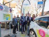 Eine Gruppe Menschen steht um eine Ladesäule und ein Elektroauto herum. Einige haben Ladekabel in der Hand.