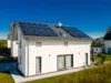 Modernes weißes Einfamilienhaus mit Satteldach und Solarpanelen, großen Fenstern und Garten, umgeben von weiteren Wohnhäusern unter klarem, blauem Himmel